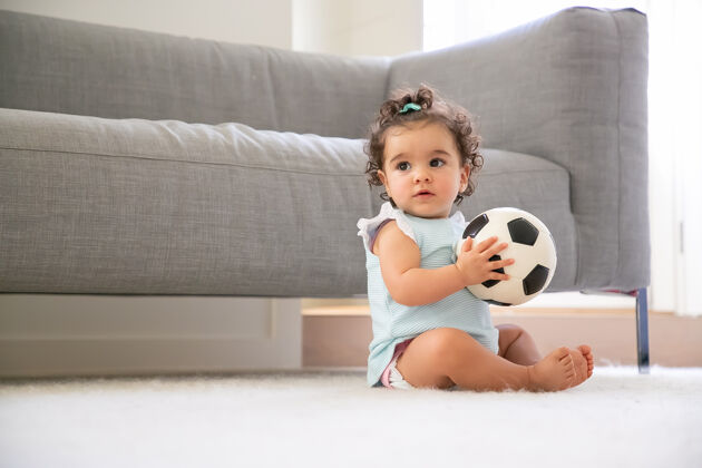 足球沉思甜美的黑发女婴 穿着淡蓝色的衣服 坐在家里的地板上 看着别处 踢足球复制空间孩子在家和童年的概念幼儿年轻地毯