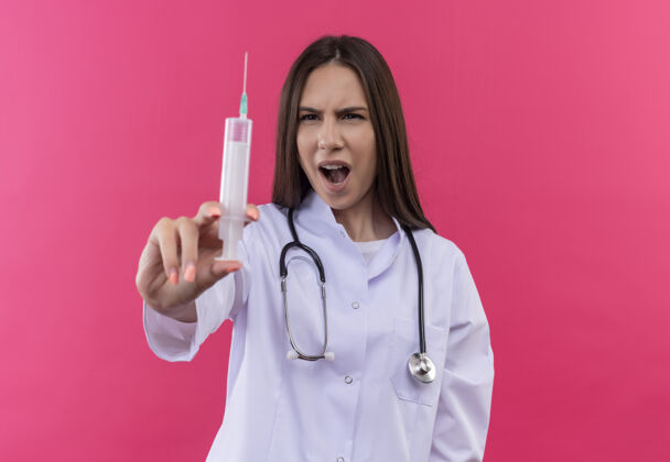 医学惊讶的年轻医生女孩穿着听诊器医用长袍 在孤立的粉红色背景下看着手中的注射器医生年轻听诊器