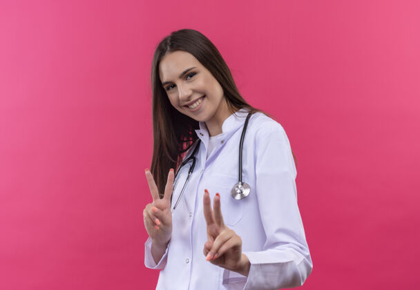 手势微笑的年轻医生女孩穿着听诊器医用长袍 在孤立的粉红色背景上显示和平姿态医疗显示穿着