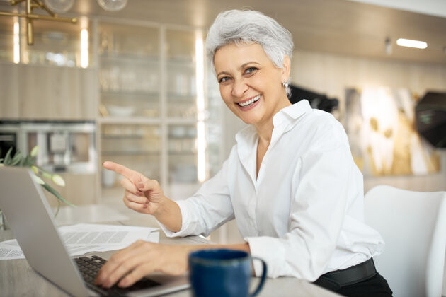 手势开朗时尚成熟的女经理在办公室工作 坐在办公桌旁 带着通用的便携式电脑 笑容灿烂 手指着食指 工作愉快创意键盘杯子