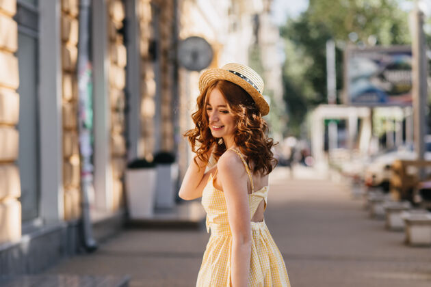 乐趣穿着黄色连衣裙的年轻女子在城市里愉快地摆着姿势戴着帽子的漂亮女孩在散步时欣赏照片的户外照片卷发波浪发成人