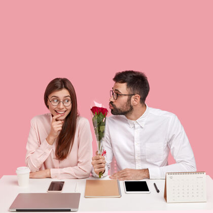 眼镜男老板爱上了年轻漂亮的同事 送上漂亮的红玫瑰 叠起嘴唇亲吻 快乐的女士收到赞美和鲜花 坐在办公室的桌面上靠着粉色的墙壁积极同事工作