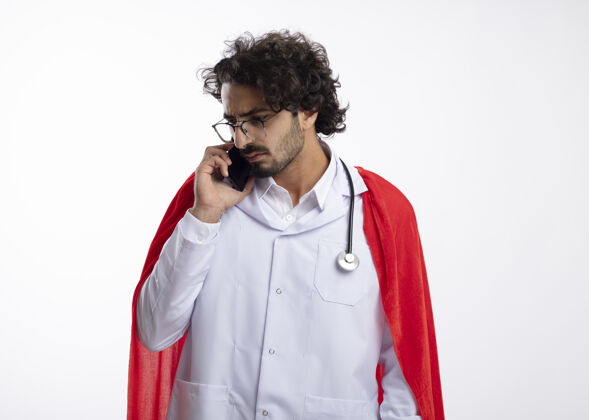 斗篷自信的年轻白种人超级英雄 戴着眼镜 身穿医生制服 披着红色斗篷 脖子上戴着听诊器 一边打电话一边看着隔离在白墙上的一边侧面穿着超级英雄