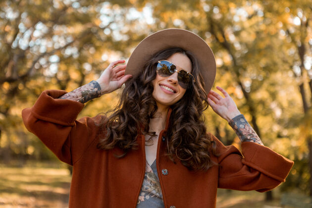 年轻迷人的时尚微笑的女人卷发漫步在街头公园穿着温暖的棕色外套秋季流行时尚 街头风格戴帽子和太阳镜魅力穿着优雅