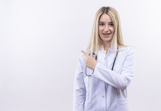 医生微笑的医生年轻的金发女孩戴着听诊器和医用长袍 戴着牙套 在孤立的白色背景上指向一边医疗微笑牙科