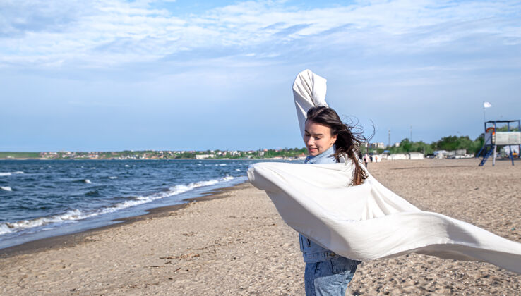 黑发海边的年轻女子在风中抱着一张大床单 享受着自由的生活方式海滩海岸夏天