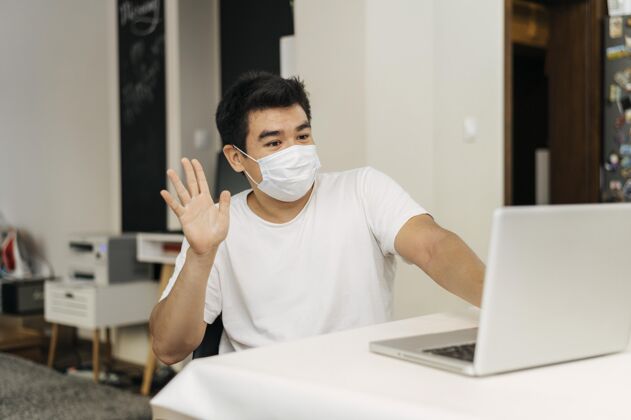 医疗面具大流行期间带着医用口罩在家的男人对着笔记本电脑挥手男性面具人