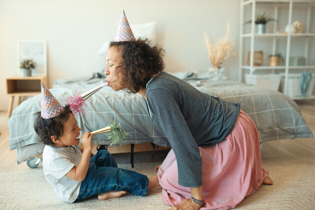 民族侧视图可爱快乐的小男孩坐在地板上 他的年轻母亲戴着锥形帽子 吹口哨帽子礼物惊喜