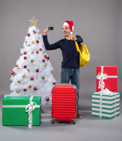 站立兴奋的年轻人拿着卡片站在圣诞树旁 送上灰色的礼物包持有购物