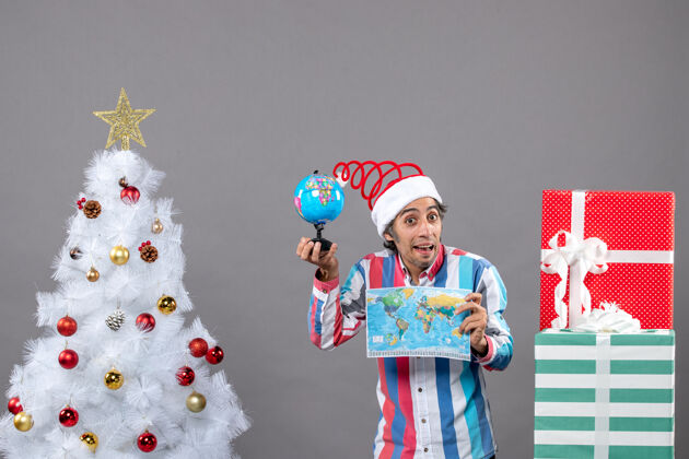 人正面图：戴着螺旋弹簧圣诞帽的年轻人拿着世界地图 在圣诞树和礼物周围展示地球年轻人圣诞礼物
