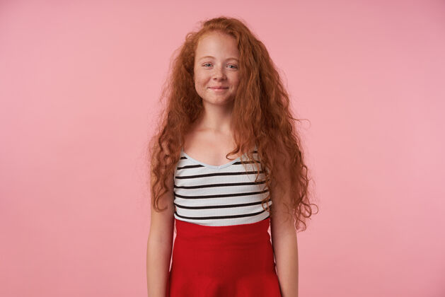 发型美丽的红头发女孩的工作室照片 长卷发摆在粉红色背景下 手放下 看着相机迷人的微笑 穿着红色裙子和条纹上衣表情红发裙子