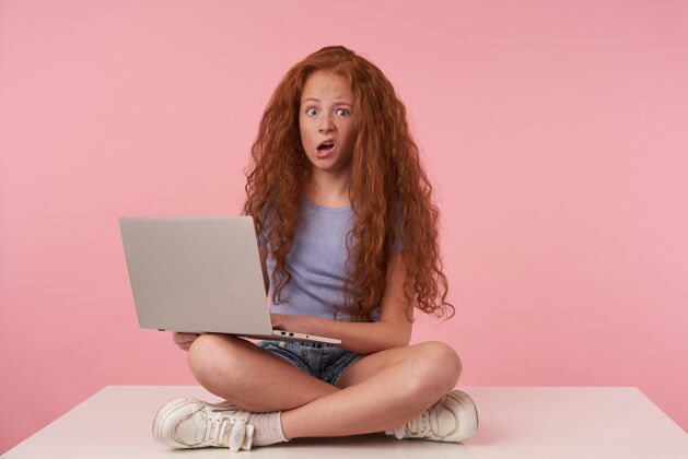 漂亮照片中 卷曲的女性少年 红色长发在粉色背景上做鬼脸 穿着休闲服交叉双腿坐着 举着笔记本电脑 睁大眼睛张大嘴巴看着相机抱着T恤运动鞋