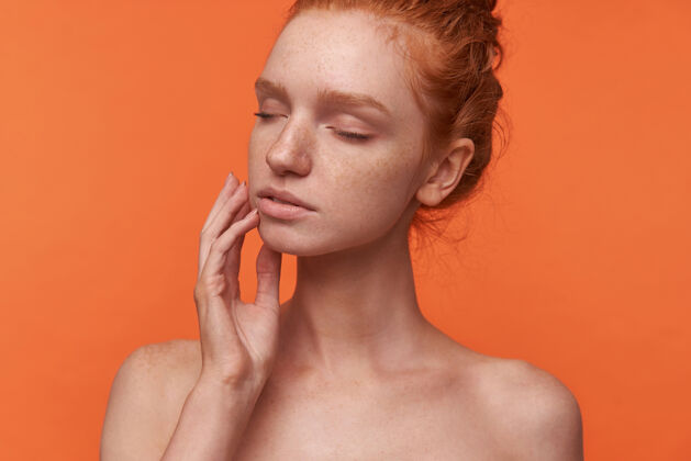 触摸摄影棚写真：年轻迷人的readhead女士留着休闲发型 闭着眼睛在橙色背景上摆姿势 用手指抚摸着她的脸优雅发髻女性
