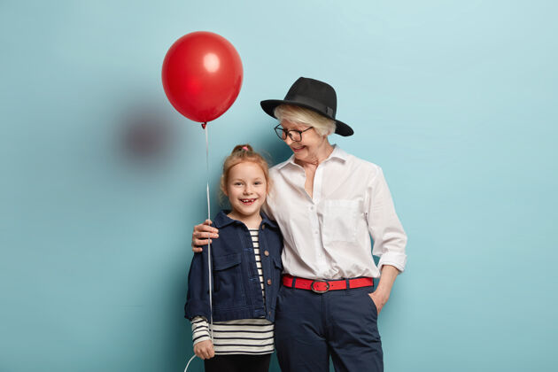 情感家庭和节日概念穿着时尚服装的爱心白发奶奶 拥抱小孙女 一起庆祝儿童节 用装饰气球站在蓝色的墙上头饰室内拥抱