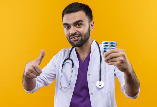 向上微笑着的年轻男医生穿着听诊器医用长袍 拿着药片 他的大拇指朝上放在孤立的黄色背景上医生年轻男性