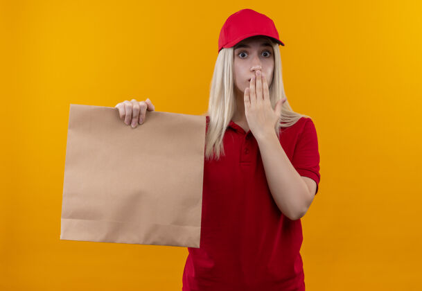 女孩一个穿着红色t恤 戴着帽子的年轻女孩拿着纸口袋 用手捂住嘴巴 背景是孤立的橙色年轻穿着嘴