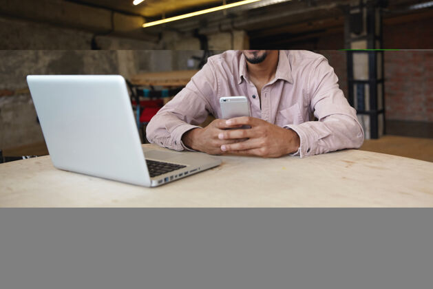 暗哑室内拍摄：英俊的黑皮肤男性 留着胡须 在同事的空间里用现代笔记本电脑远程工作 用智能手机和伙伴视频聊天 对着镜头开心地微笑摆姿势成人漂亮