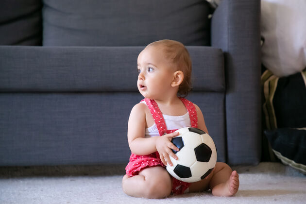 周末可爱的女婴拿着足球 光着脚坐在地毯上 看着别处可爱的婴儿穿着红色的工作服短裤独自在家里玩耍假日 周末和童年的概念幼儿沙发工作服