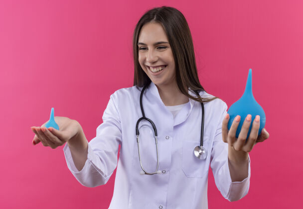 看带着微笑的年轻医生女孩穿着听诊器医用长袍 在孤立的粉色背景下看着手里的灌肠液穿着医学手