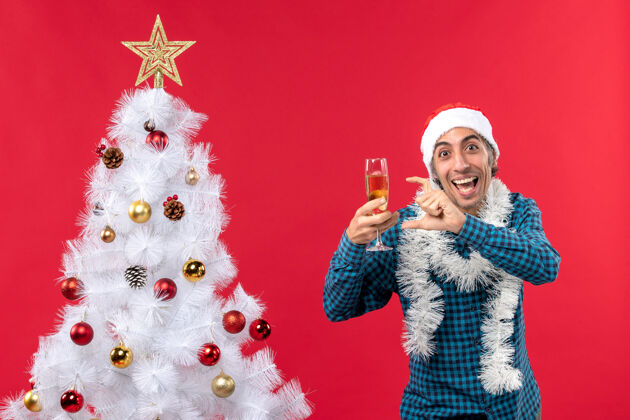 指向圣诞节心情与情绪激动的年轻人圣诞老人帽子在一个蓝色的衬衫剥离举行一杯葡萄酒指着圣诞树附近的东西年轻人庆祝帽子