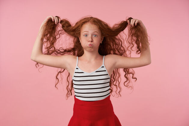 手一幅长发卷发女孩的画像 她一边玩着自己的红发 一边站在粉色背景上 鼓起脸颊开玩笑 扬起眉毛看着相机积极红发制造