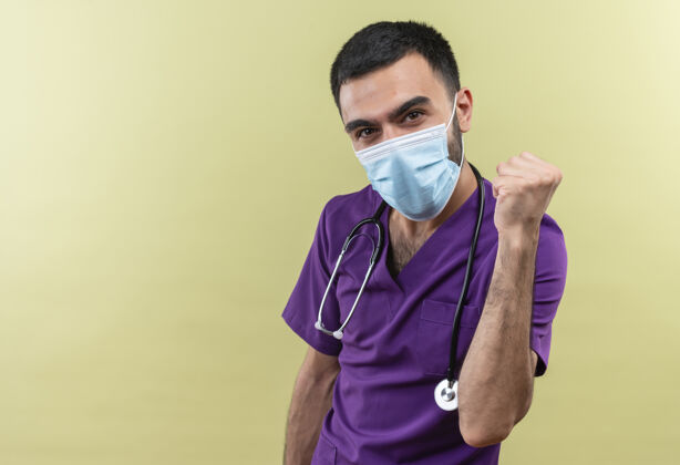 穿着年轻的男医生穿着紫色的手术服 戴着听诊器医用口罩 在绿色的背景上表示同意男性手势衣服