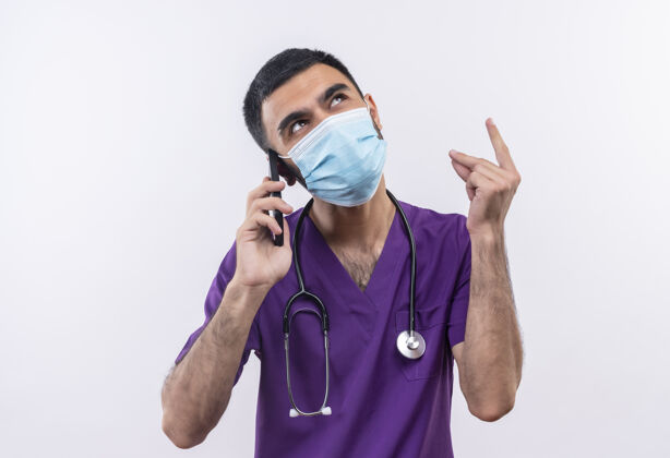 男性看着身穿紫色手术服和听诊器医用面罩的年轻男医生在孤立的白色背景下讲电话戴外科医生年轻