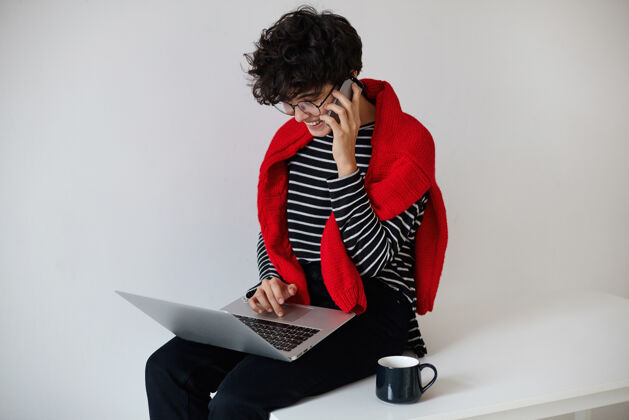 女士积极的年轻漂亮的短发卷曲的黑发女人坐在她的腿上的笔记本电脑和电话交谈 而在白色的背景摆姿势通话交谈针织