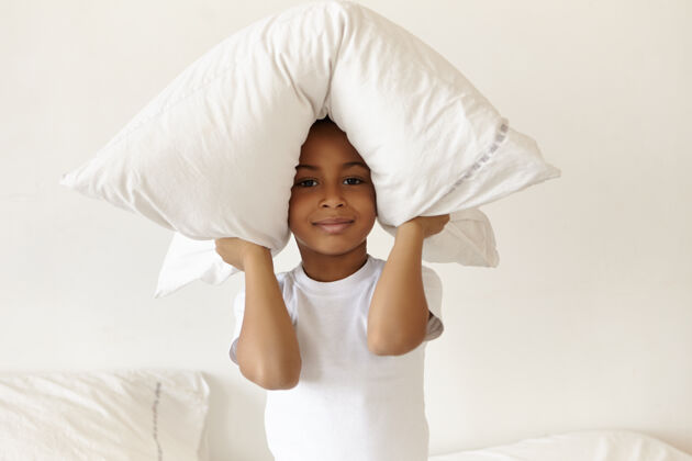 枕头横向拍摄的可爱快乐的非洲裔美国小男孩穿着睡衣坐在卧室里游戏早晨柔软
