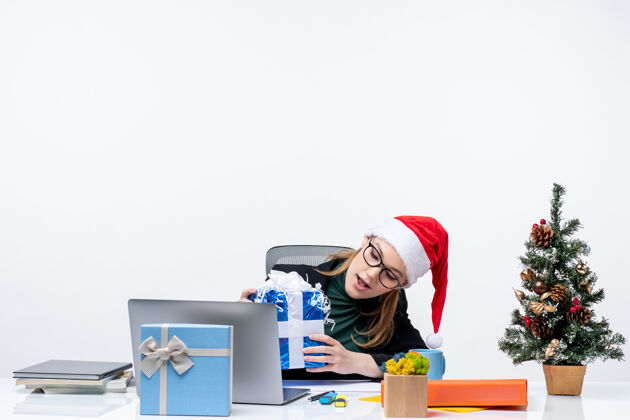 人圣诞气氛与戴着圣诞老人帽子和眼镜的年轻女子坐在一张桌子上看着她的礼物令人惊讶的白色背景桌子年轻人帽子