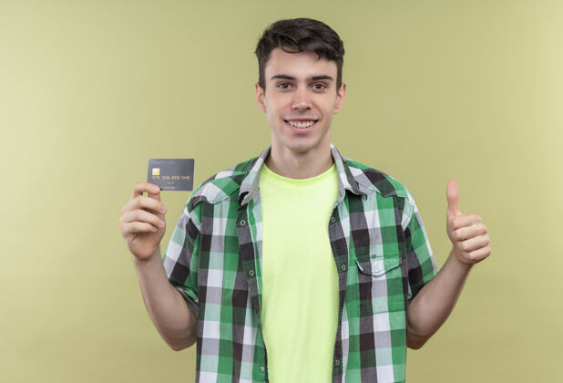 拇指微笑的白人年轻人穿着绿色衬衫 举着信用卡 在孤立的绿色背景上竖起大拇指拿着微笑向上