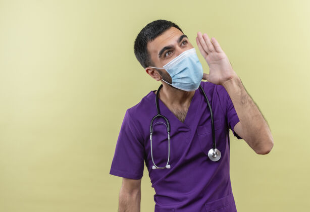 年轻看着身边年轻的男医生穿着紫色的手术服 戴着听诊器医用面罩 在孤立的绿色背景上呼唤着某人看绿色外科医生