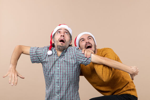 伙计们前视图两个戴着圣诞帽的吓坏了的家伙 一个躲着另一个 同时在米色孤立的背景下看着高处米色男人藏起来