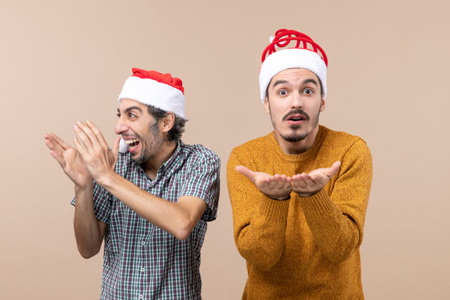 男性正面图两个戴着圣诞帽的男人站在米色孤立的背景上 一个拍手 另一个张开双手手伙计们鼓掌