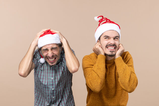 站着正面图两个戴着圣诞帽的吓坏了的家伙 一个把手放在头上 另一个放在耳朵上 站在米色的孤立背景上前面圣诞老人微笑