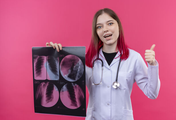 长袍微笑着的年轻医生女孩穿着听诊器医用长袍 举着x光片 拇指朝上放在粉红色的孤立背景上粉红年轻医生
