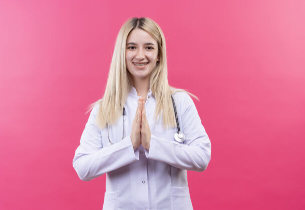 医生微笑的医生年轻的金发女孩穿着医用长袍戴着听诊器和牙套 在孤立的粉色背景上显示祈祷的手势听诊器女孩手势