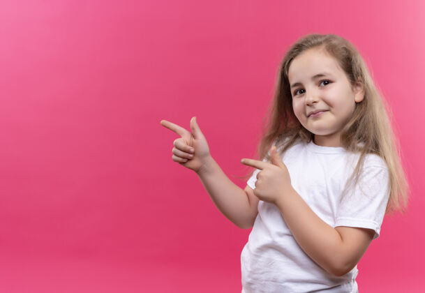 小微笑的小女孩穿着白色t恤指向侧面 背景是孤立的粉色点学校穿