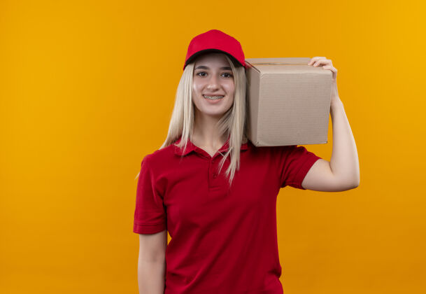 牙科微笑的小女孩穿着红色t恤 戴着帽子 戴着牙套 肩上扛着一个盒子 背景是孤立的橙色抱着戴着年轻