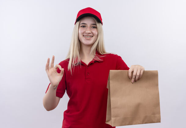 支架微笑的小女孩穿着红色t恤 戴着帽子 戴着牙套 拿着纸袋 在孤立的白色背景上显示出良好的姿态口袋送货穿着