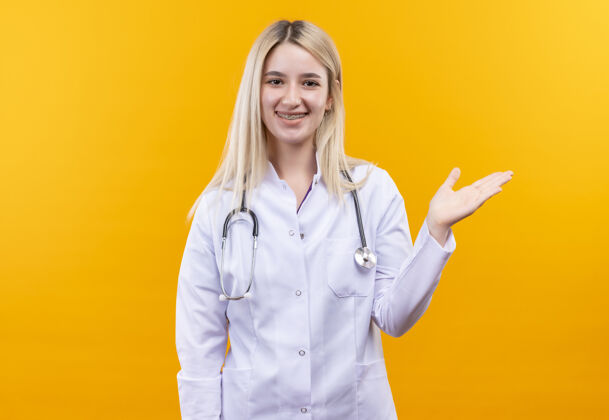 长袍微笑的医生穿着医用长袍戴着听诊器和牙套的年轻女孩指着隔离的黄色背景的侧面穿年轻医疗