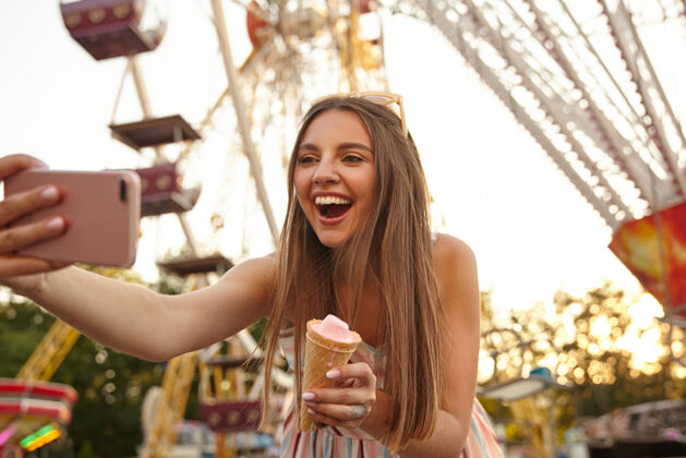 温暖快乐迷人的年轻黑发女性的肖像 穿着夏装 用智能手机在户外自拍 手里拿着冰激凌筒 快乐地微笑衣服公园微笑