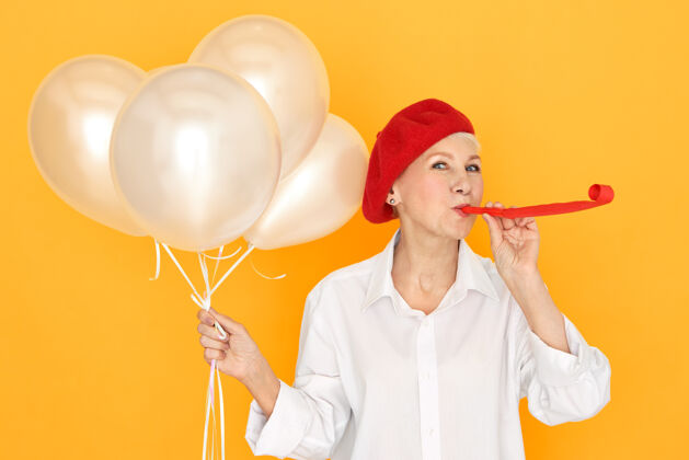 时尚一位戴着红帽子的退休妇女 享受着聚会 招待她的孙子孙女吹着口哨 手里拿着白色的氦气球生日角举行