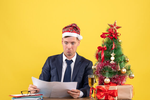 会议年轻的商人坐在圣诞树旁的桌子旁看报纸 黄色的礼物摆在桌子上圣诞树礼物支票