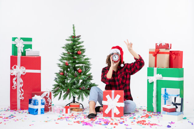 微笑前视图的年轻女子坐在圣诞礼物周围拿着白色墙上的时钟人快乐节日