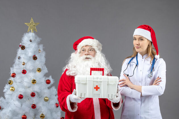 圣诞老人圣诞老人和给他急救箱的女医生在灰色墙上的正视图援助圣诞老人快乐