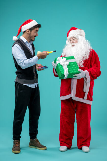 男人前视图：圣诞老人和正在蓝色背景上做笔记的年轻男性音乐家圣诞老人采取