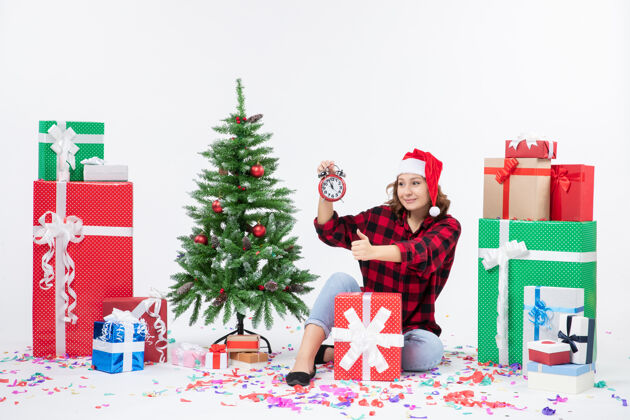 前面前视图的年轻女子坐在圣诞礼物周围拿着白色墙上的时钟人礼物时钟