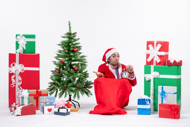 时钟惊讶的年轻人坐在地上庆祝新年或圣诞节 拿着钟靠近礼物和装饰圣诞树礼物年轻人礼物