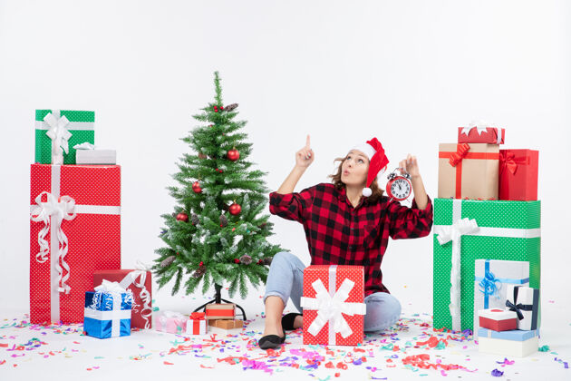 礼物前视图的年轻女子坐在圣诞礼物周围拿着白色墙上的时钟快乐快乐礼物